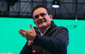 Hombre de mediana edad, con cabello oscuro y rizado, en chaqueta oscura y camisa blanca, habla con micrófono en mano frente a fondo verde.