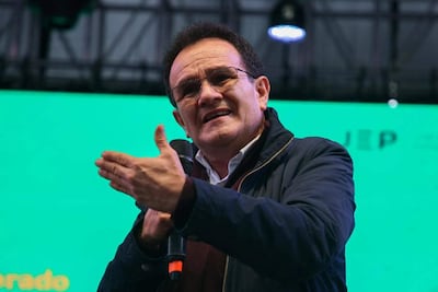 Hombre de mediana edad, con cabello oscuro y rizado, en chaqueta oscura y camisa blanca, habla con micrófono en mano frente a fondo verde.