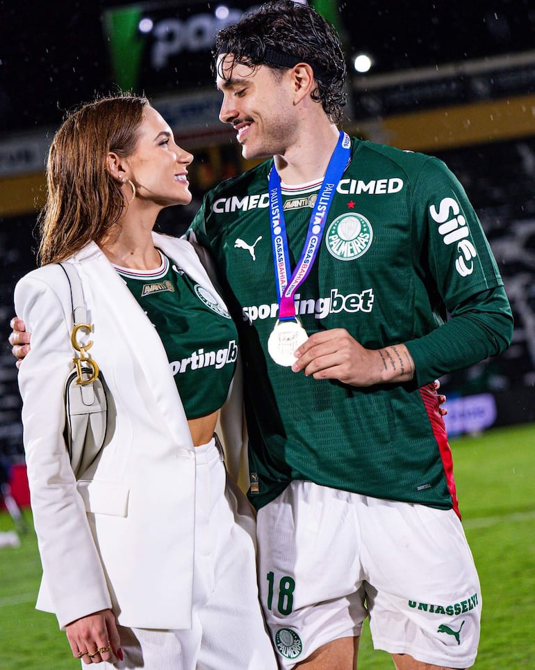 Mujer sonriente en traje blanco junto a hombre con camiseta verde de Palmeiras, ambos en ambiente festivo tras un partido.