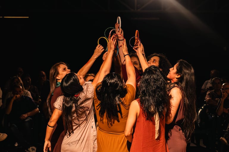 Diez mujeres en semicírculo levantan aros coloridos, con expresiones de alegría en un auditorio con audiencia presente.