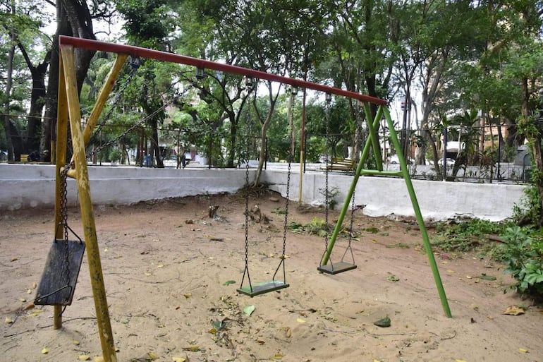 Columpio metálico rojo y verde en un parque infantil vacío rodeado de árboles, con arena y hojas caídas en el suelo.