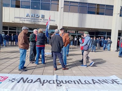 Grupo de adultos mayores, una mujer en chaleco negro habla con un hombre canoso. Cartel con hashtag '#NoApaguenLaANDE' en el suelo.