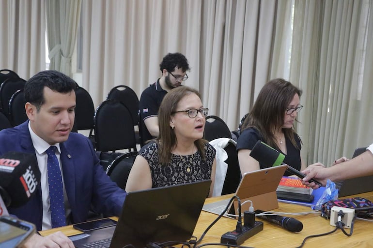 Christian Benítez en chaqueta azul y Patricia Sánchez en blusa negra, ambos con expresión de sorpresa en mesa rodeada de dispositivos.