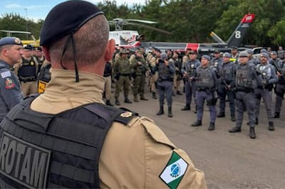 Oficial de policía con chaleco 'ROTAM' observa a soldados en formación, en un ambiente serio y militarizado, con helicóptero al fondo.
