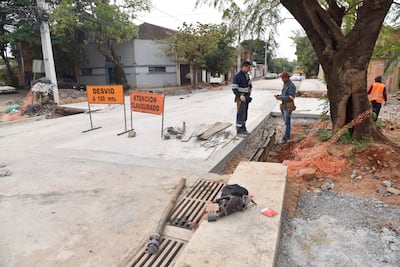 Dos trabajadores, uno en uniforme oscuro y otro con gorra roja, supervisan la obra cerca de una trinchera abierta en una calle urbana.