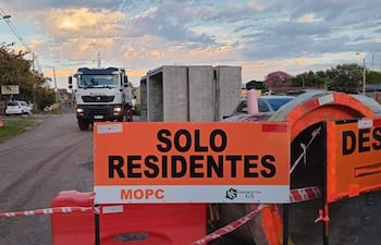 Cartel de 'SOLO RESIDENTES' en construcción vial, con casas y un camión en el fondo bajo un cielo nublado.