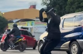 Persona con traje negro, camisa blanca y sombrero bailando en una calle con vehículos de fondo.