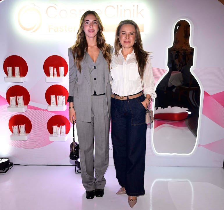 Dos mujeres sonrientes visten un traje gris y camisa blanca con jeans, posando frente a un fondo decorativo con productos cosméticos.