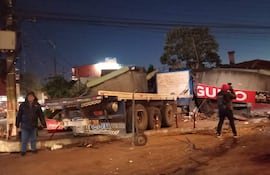 Hombre en abrigo oscuro y pantalones claros observa un camión con daños. El entorno muestra signos de desorden.