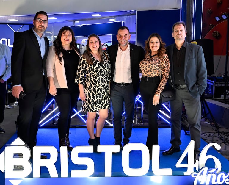 Grupo de seis personas sonriendo, tres hombres con traje y corbata, tres mujeres con ropa de moda, fondo con cartel luminoso de Bristol 46.