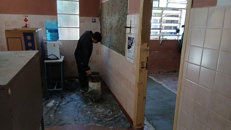 Hombre vestido con ropa oscura agachado, aplicando mezcla en la pared con cubos de pintura en un espacio de construcción.
