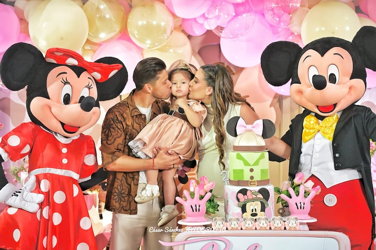 Pareja sonriente con niño en brazos, rodeados de globos pasteles y pastel de Minnie Mouse.