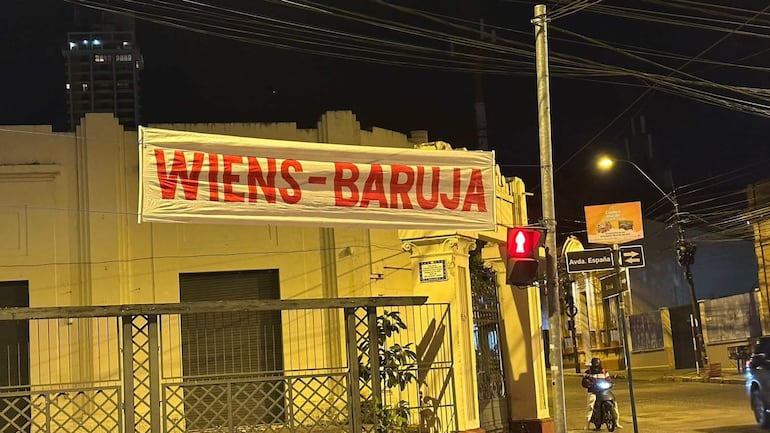 Pasacalles iluminados que dicen 'WIENS-BARUJA' en una calle desierta, con semáforo en rojo y edificios antiguos.
