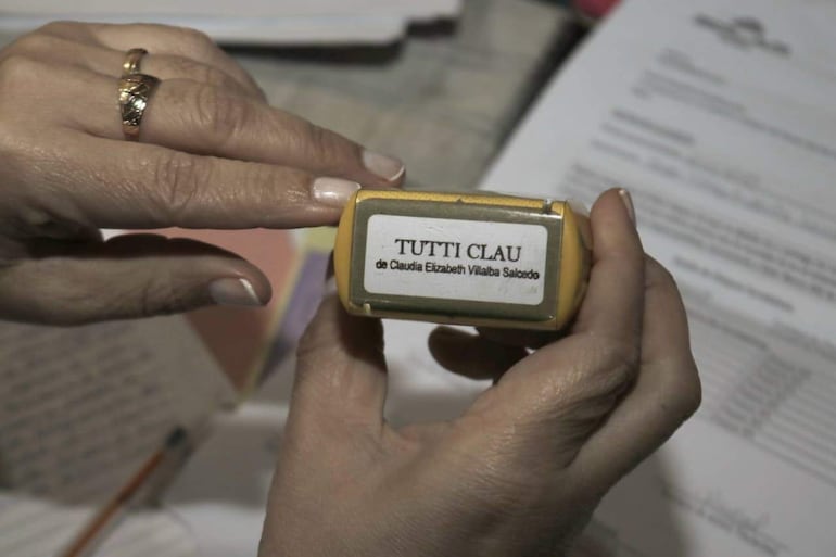 Sello amarillo con insignia 'TUTTI CLAU' y nombre de Claudia Elizabeth Villalba Salcedo, sostenido por una mano en un entorno de trabajo.
