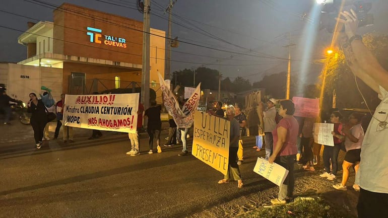 Vecinos sostienen carteles de protesta, iluminados por farolas, en una calle oscura, pidiendo ayuda al MOPC.