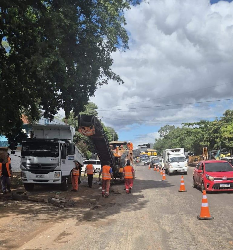 Siete trabajadores con chalecos naranja operan maquinaria en una carretera de cielo nublado, rodeados de conos de señalización y vehículos en movimiento.