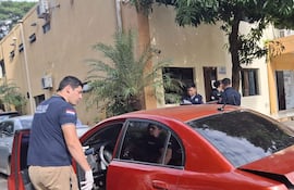 Hombre con camiseta de 'Policía' inspecciona auto rojo mientras otro está dentro; ambiente urbano con oficinas y plantas visibles.