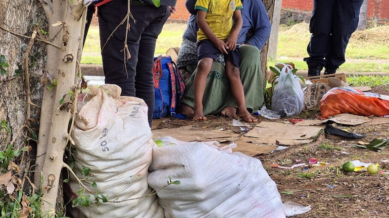 Un niño con camiseta amarilla está sentado junto a dos adultos en un ambiente sucio, lleno de cartones y desechos.