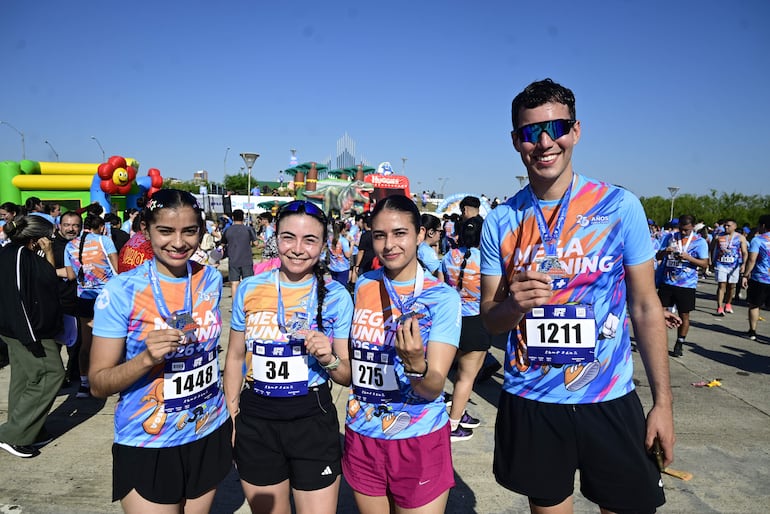 Cuatro corredores con camisetas azules y naranjas sonríen, sosteniendo medallas en un ambiente festivo en la Costanera.