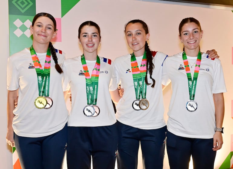 Cuatro mujeres sonríen, vestidas con camisetas blancas, luciendo medallas en ambiente colorido y festivo.