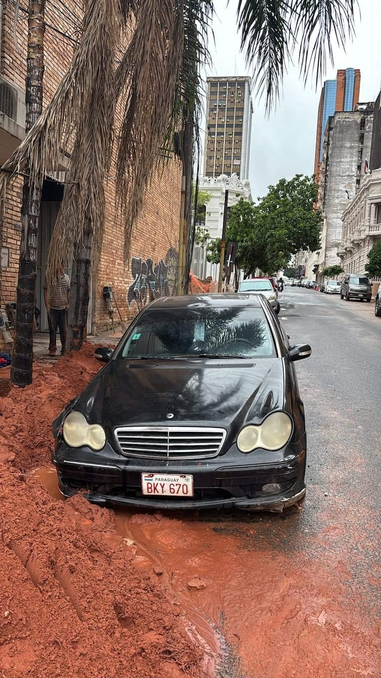 Hombre de pie observando su Mercedes Benz negro cubierto de tierra, con edificios y palmeras al fondo bajo un cielo nublado.