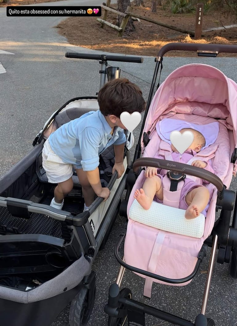 Niño pequeño con camiseta clara e inclinándose hacia su hermana bebé en cochecito rosa, en un parque con árboles.