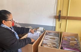 Mujer de cabello recogido en coleta, con chaqueta negra y bufanda, organiza documentos en cajas de madera.
