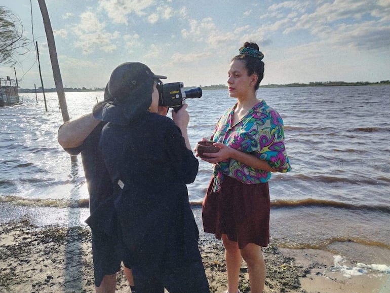 Mujer con blusa colorida y falda oscura, sosteniendo un objeto, mientras otra persona graba la escena en la playa.
