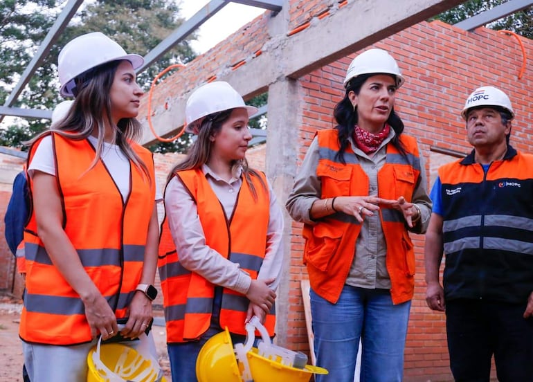Hugo Arce y Claudia Centurión con cascos y chalecos en obra, Claudia gesticula mientras dos mujeres la escuchan con atención.