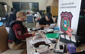 Mujer con chaleco antibalas escribe en un cuaderno, mientras hombre toma notas sobre documentos en un ambiente policial.