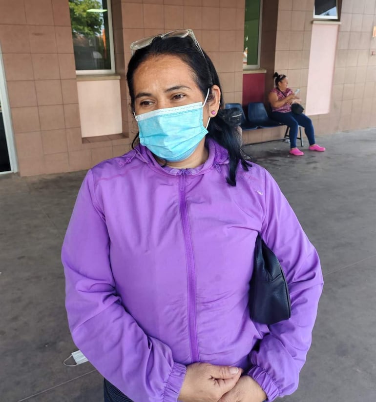 Mujer de pie con chaqueta morada y mascarilla azul, dos amigas la acompañan en un ambiente público con sillas y cerámica.