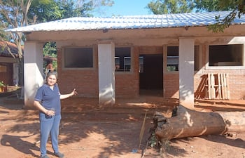 Mujer de pie frente a construcción, con cabello largo, camiseta oscura y vaso en mano, señalando la obra en un entorno rural.