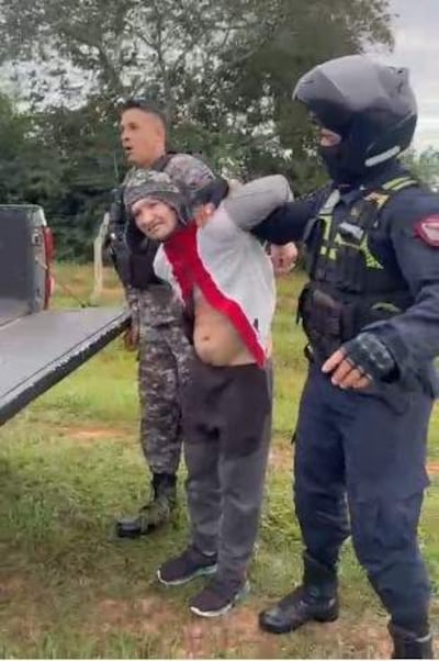 Individuo con camiseta blanca y roja, detenido por un policía en traje negro, visiblemente incómodo en un área rural.