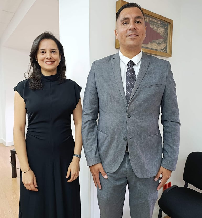 Mujer de cabello oscuro y vestido negro sonríe al lado de un hombre con traje gris claro y corbata con puntos oscuros.