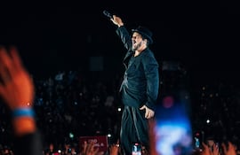 Ricardo Arjona en el escenario, con saco a rayas y sombrero negro, sosteniendo un micrófono, mientras el público aplaude y levanta manos.