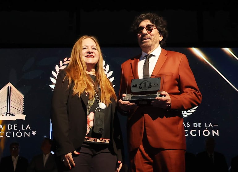 Mujer sonriente de blusa negra y hombre con traje marrón sostienen un trofeo en un evento formal.