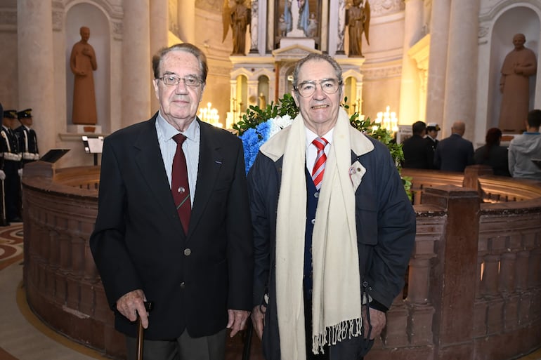 Dos hombres bien vestidos, uno en traje negro y corbata roja, y otro en abrigo azul y bufanda, frente a decoración floral.