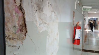 Pasillo deteriorado con paredes descascaradas y manchas de humedad. Persona con abrigo negro al fondo, extintor en la pared.
