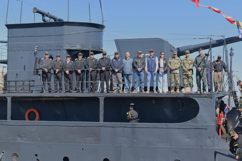Grupo de personas en la cubierta de un buque militar con uniforme de marina y ambiente solemne en un día despejado.