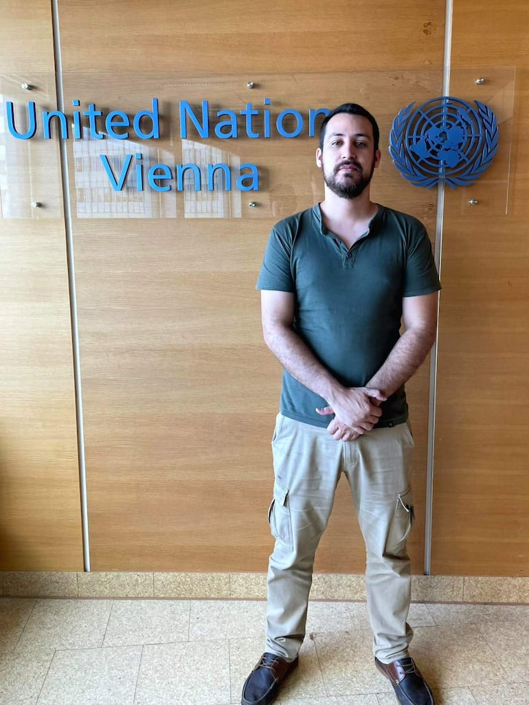 Lic. Alcides Maximiliano Caballero vestido con camiseta verde y pantalones claros, de pie frente al logotipo de la ONU en un ambiente institucional.