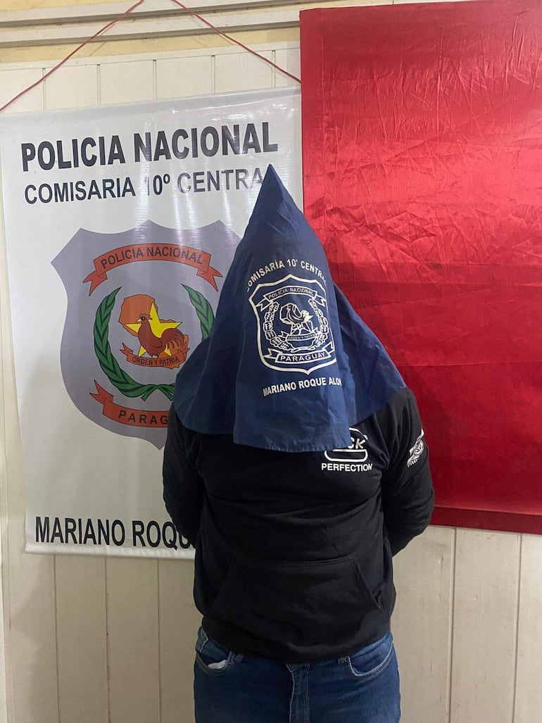 Hombre de espaldas con sudadera negra y pañuelo azul frente a un cartel de la Comisaría 10° Central en Paraguay.