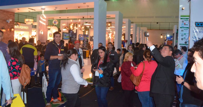 Mujer de abrigo oscuro conversando con otra persona, luces modernas decoran un evento lleno de gente.