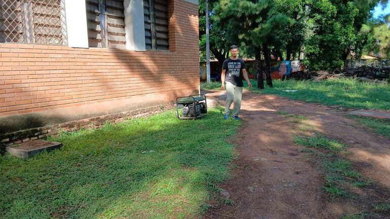 Hombre con camiseta negra y pantalones claros camina hacia un generador eléctrico en el césped del patio del colegio.