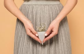 Persona con blusa amarilla y falda gris sostiene un copo menstrual blanco con flores, sobre fondo naranja en un ambiente cálido.