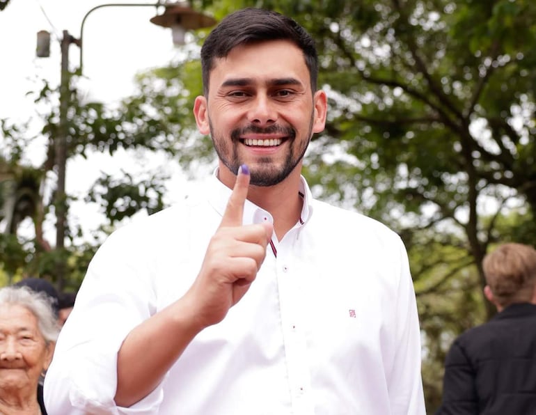 Hombre sonriente con camisa blanca levanta un dedo índice, rodeado de personas en un ambiente natural.