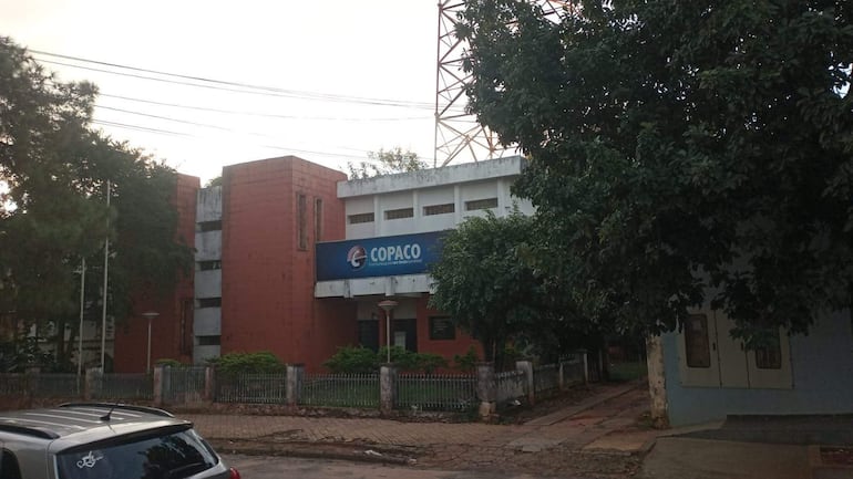 Edificio de COPACO en Caaguazú, sin personas visibles, rodeado de árboles y arbustos, con diseño simple y paredes rojizas.
