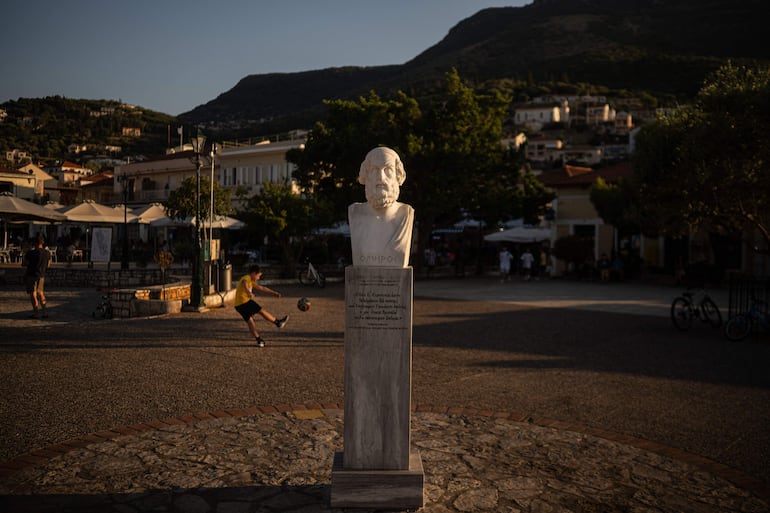 Un niño juega al fútbol junto a un busto de mármol del antiguo poeta griego Homero, en la plaza central de Vathí, en la isla de Ítaca, en el mar Jónico, el 3 de agosto de 2026.