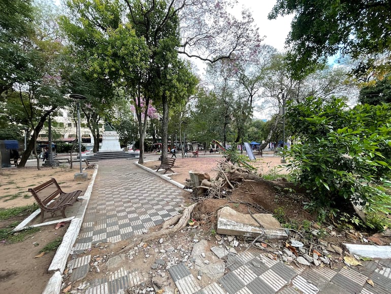 Parque urbano sin personas, monumento en el centro, sendero rodeado de árboles y bancos metálicos en un día nublado.