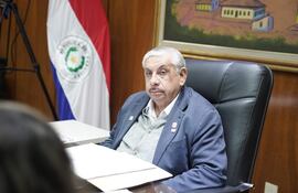Isaías Ricardo Fretes en chaqueta azul y suéter gris, sentado en oficina con bandera de Paraguay y documentos sobre la mesa.