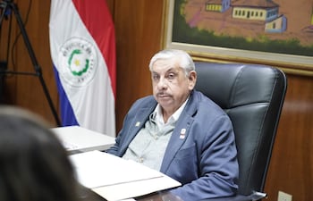 Isaías Ricardo Fretes en chaqueta azul y suéter gris, sentado en oficina con bandera de Paraguay y documentos sobre la mesa.